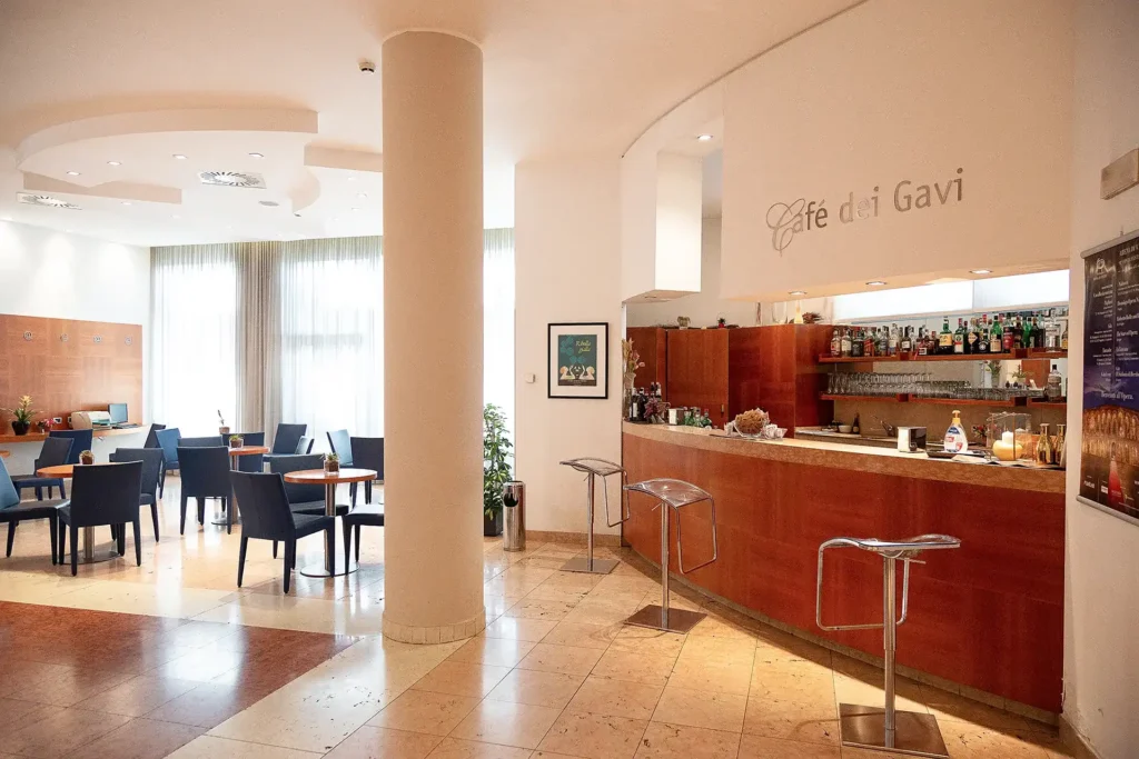 Nell'immagine, il Lounge Bar dell'Hotel Fiera Verona: si vede il bancone del bar sulla destra, con degli sgabelli. Sulla sinistra, invece, ci sono alcuni tavolini tondi con delle sedie.